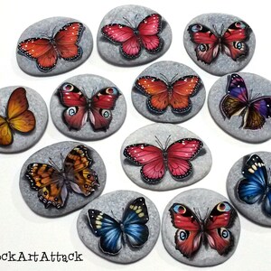 May include: Hand-painted butterfly art on smooth, gray stones. Each stone features a unique butterfly design in vibrant colors like red, orange, blue, and purple. The artwork is detailed and realistic, showcasing the beauty of nature.