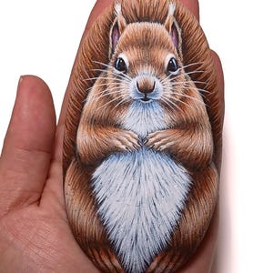 May include: A hand-painted rock shaped like a squirrel. The squirrel has brown fur, a white chest, and a fluffy tail. The rock art is detailed, with visible whiskers and a cute expression. The artwork is held in a person's hand.