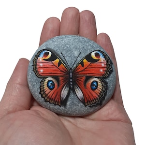 May include: A hand-painted, gray stone features a vibrant butterfly design. The butterfly has red, orange, yellow, and blue details, with intricate wing patterns. The stone is smooth and round, showcasing the artwork.