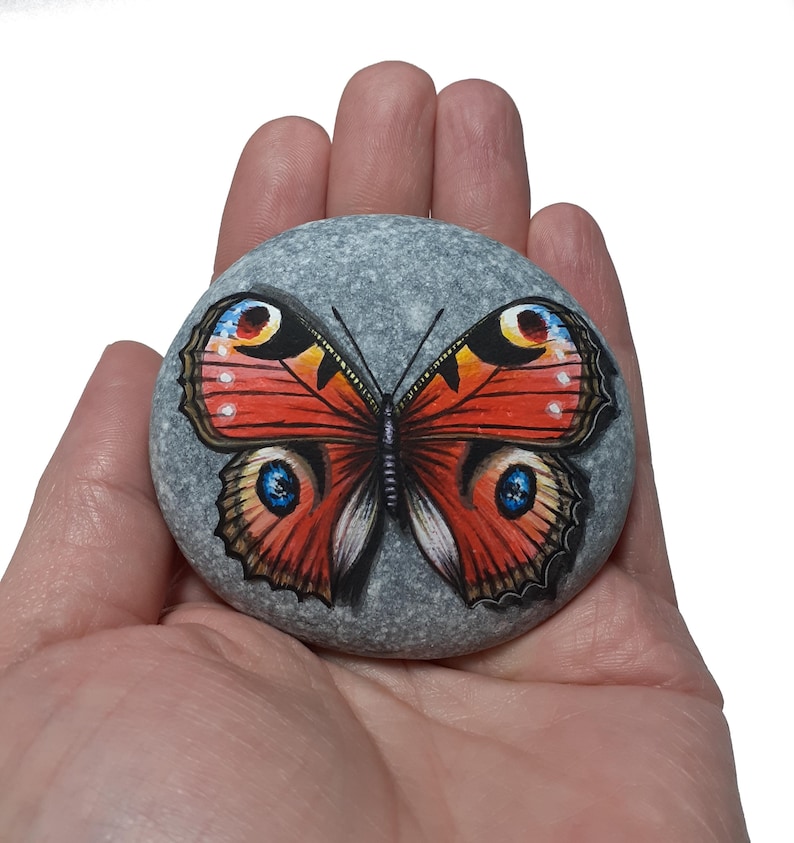 May include: A hand-painted, gray stone features a vibrant butterfly design. The butterfly has red, orange, yellow, and blue details, with intricate wing patterns. The stone is smooth and round, showcasing the artwork.