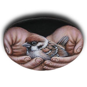 May include: A hand-painted oval stone featuring a detailed illustration of a small brown and white sparrow nestled within two cupped hands. The background of the painting is solid black, contrasting with the realistic textures of the bird's feathers and the skin tones of the hands depicted on the smooth, rounded surface.