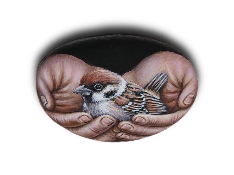 May include: A hand-painted oval stone featuring a detailed illustration of a small brown and white sparrow nestled within two cupped hands. The background of the painting is solid black, contrasting with the realistic textures of the bird's feathers and the skin tones of the hands depicted on the smooth, rounded surface.