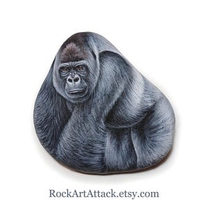 May include: A smooth, rounded stone featuring a detailed hand-painted portrait of a silverback gorilla. The gorilla is depicted in shades of black, gray, and white, with focused brown eyes and textured fur. The text RockArtAttack.etsy.com is printed below.