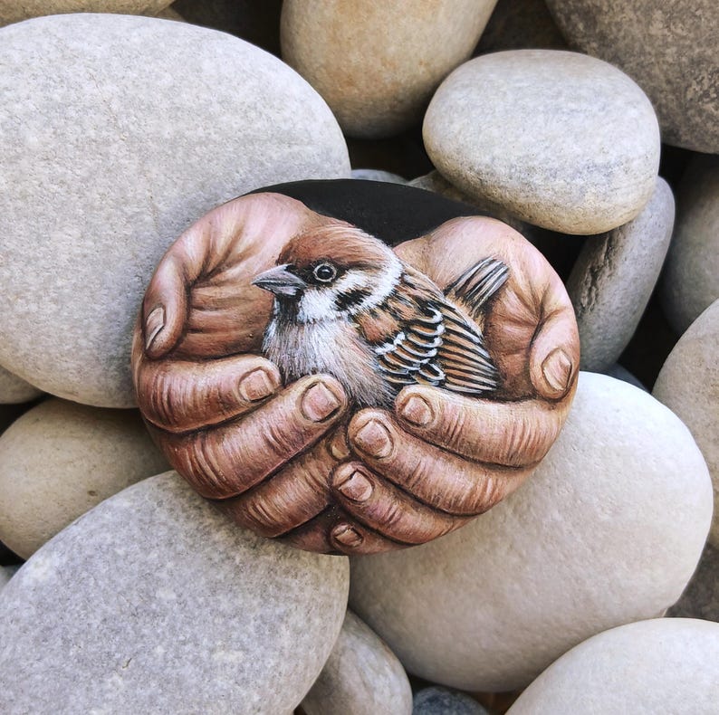 May include: A smooth, rounded stone featuring a detailed, realistic painting of a small brown and white sparrow nestled within two cupped palms. The artwork captures the intricate feather textures of the bird and the skin details of the hands.