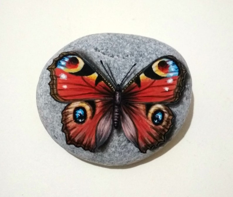 May include: A hand-painted stone featuring a vibrant butterfly design. The butterfly has red wings with blue, yellow, and black accents. The stone is gray and oval-shaped, showcasing a nature-inspired art piece.