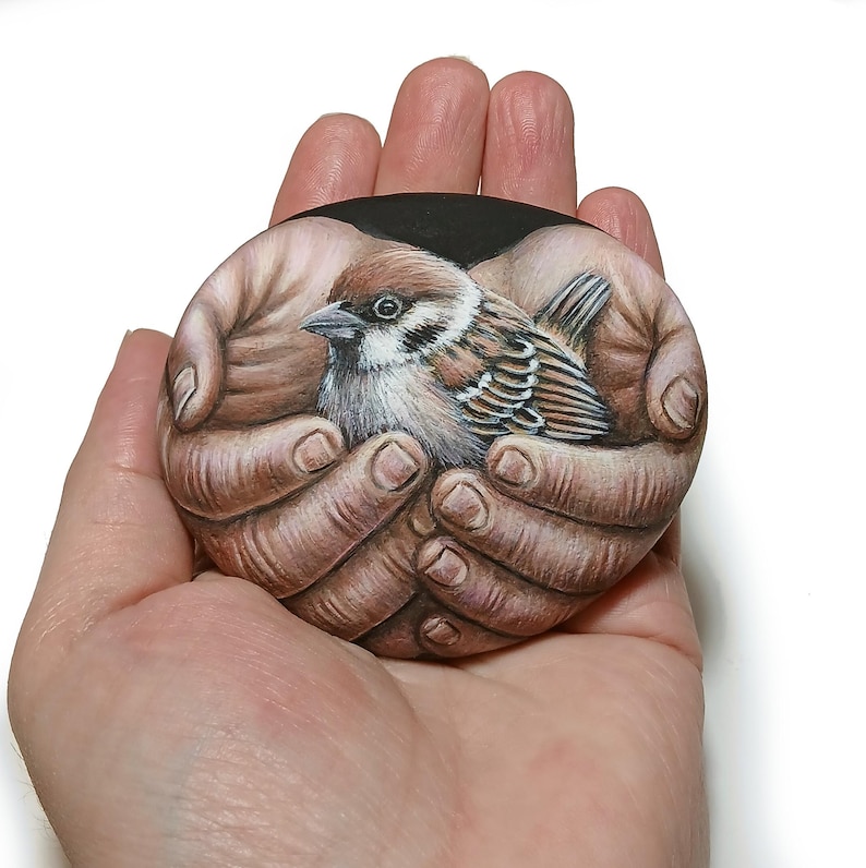 May include: A smooth, rounded stone featuring a detailed, realistic painting of a small brown and white sparrow nestled within two cupped hands. The artwork uses shades of brown, tan, white, and black to depict the bird and the skin texture.