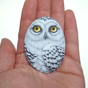 Peut inclure: Une pierre de forme ovale peinte avec un harfang des neiges détaillé. Le hibou présente des plumes blanches et grises, un bec noir pointu et deux grands yeux jaune vif. La peinture recouvre toute la surface de la pierre lisse et arrondie.