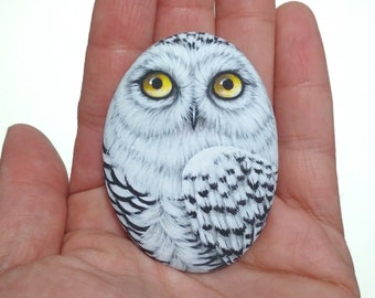 May include: An oval-shaped stone painted with a detailed snowy owl. The owl features white and grey feathers, a sharp black beak, and two large, bright yellow eyes. The painting covers the entire surface of the smooth, rounded stone.