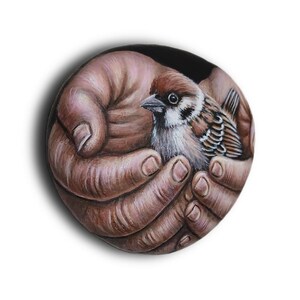 May include: A circular painted stone featuring a detailed illustration of two cupped hands holding a small brown and white sparrow. The bird has distinct dark markings around its eyes and beak, while the hands are rendered in warm brown tones.