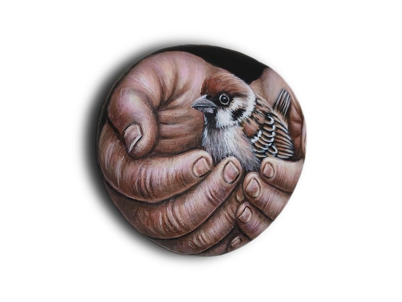 May include: A circular painted stone featuring a detailed illustration of two cupped hands holding a small brown and white sparrow. The bird has distinct dark markings around its eyes and beak, while the hands are rendered in warm brown tones.