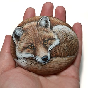 May include: A smooth, oval-shaped river stone featuring a detailed hand-painted illustration of a curled-up red fox. The fox has golden-brown eyes, a black nose, and white fur accents around its ears, cheeks, and chest, set against a brown fur.