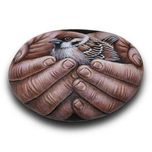 May include: A smooth, oval-shaped stone featuring a detailed, realistic painting of a small brown and white sparrow resting within a pair of cupped hands. The artwork uses natural earth tones to depict the bird's feathers and the skin texture.