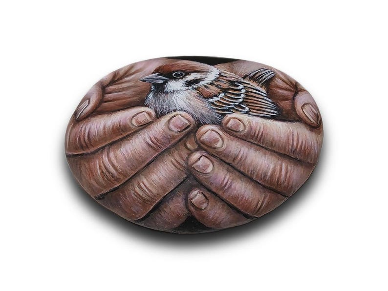 May include: A smooth, oval-shaped stone featuring a detailed, realistic painting of a small brown and white sparrow resting within a pair of cupped hands. The artwork uses natural earth tones to depict the bird's feathers and the skin texture.