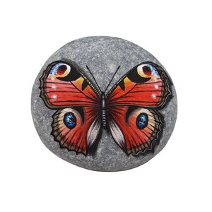 May include: A hand-painted, gray stone featuring a vibrant butterfly design. The butterfly has red, orange, and black wings with blue and yellow accents. The stone is oval-shaped and sits against a white background.