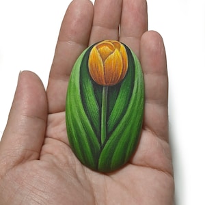 May include: An oval-shaped stone featuring a hand-painted illustration of a single yellow and orange tulip. The flower is depicted with a central stem and several long, pointed green leaves that curve upward, framing the bloom against a dark background.