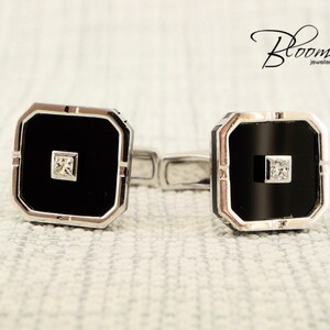 May include: A pair of square cufflinks with a black center and a small square diamond. The cufflinks have a silver-colored border and are connected by a silver bar. The Bloom Jewellery logo is in the upper right corner.