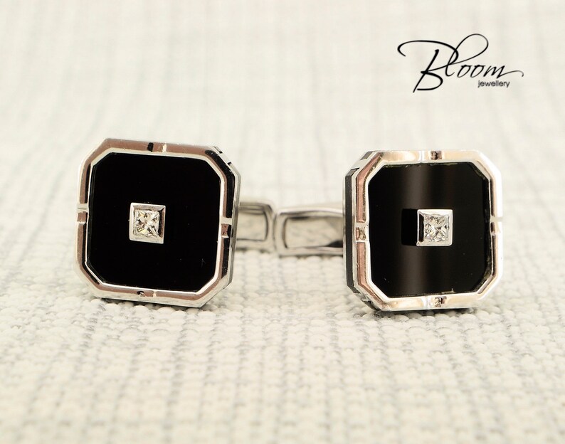 May include: A pair of square cufflinks with a black center and a small square diamond. The cufflinks have a silver-colored border and are connected by a silver bar. The Bloom Jewellery logo is in the upper right corner.
