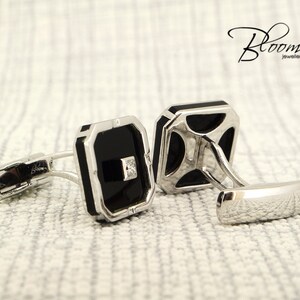 May include: A pair of silver cufflinks with black rectangular insets and a small square diamond. The cufflinks have a polished finish and are set against a textured, off-white background. The Bloom Jewellery logo is visible.