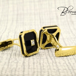 May include: A pair of gold-toned cufflinks with black square insets and a small diamond accent. The cufflinks have a rectangular shape with rounded corners and are displayed on a textured surface. The Bloom Jewellery logo is visible.