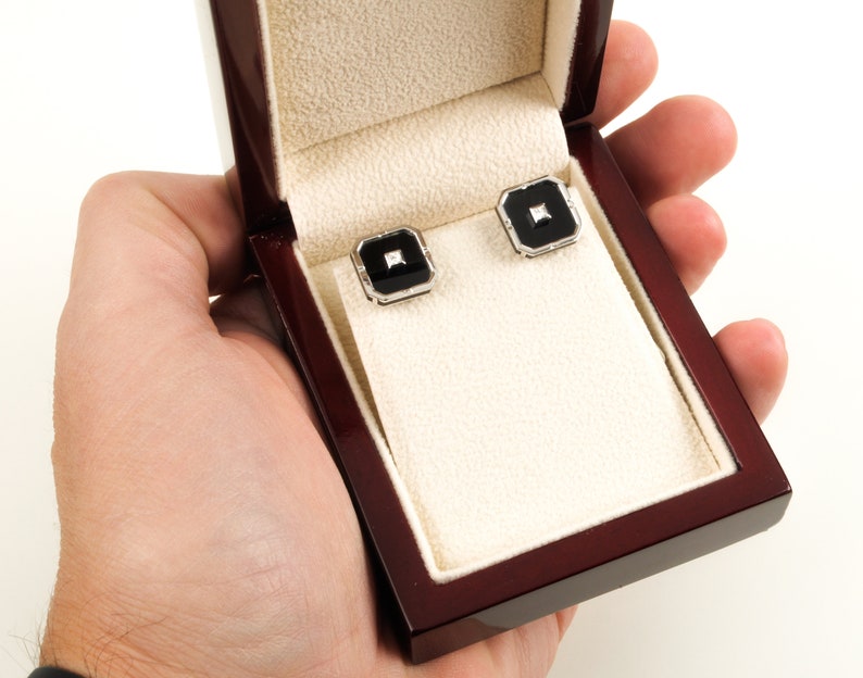 May include: A pair of square cufflinks with black onyx centers and a small diamond accent, set in a silver-tone metal. The cufflinks are displayed in an open, polished wooden box with a cream-colored interior.