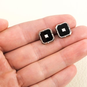May include: A pair of square cufflinks with a black center and a small, square, clear stone in the middle. The cufflinks have a silver-colored border. The cufflinks are displayed on a light-colored surface.