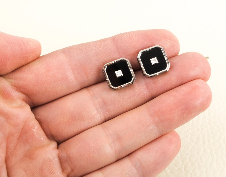 May include: A pair of square cufflinks with a black center and a small, square, clear stone in the middle. The cufflinks have a silver-colored border. The cufflinks are displayed on a light-colored surface.