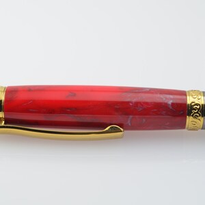 May include: A red and white marbled pen with gold accents and a black metal clip. The pen has a gold band with an engraved design.