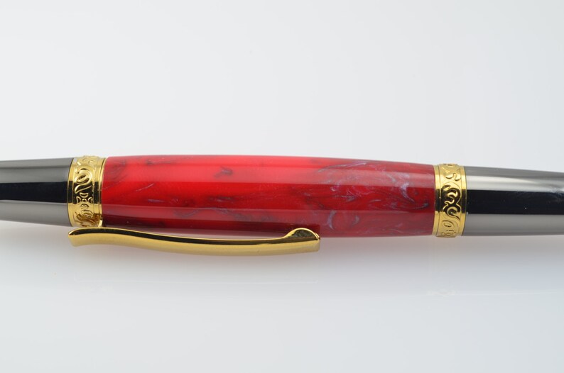May include: A red and white marbled pen with gold accents and a black metal clip. The pen has a gold band with an engraved design.