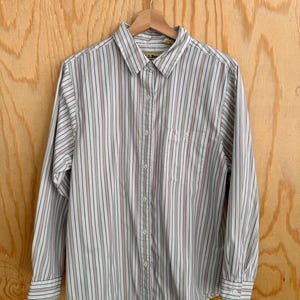 May include: A button-down shirt with vertical stripes in white, navy, and burgundy. The long-sleeved shirt has a chest pocket and is hanging on a wooden hanger. The shirt is made of a lightweight fabric.