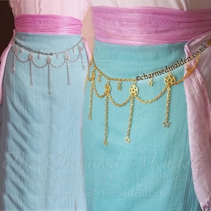 May include: Two decorative waist chains, one silver and one gold, each with dangling chains and star and pentagram charms. The chains are displayed against a light blue fabric, with a pink sash above. The text "The Charmed Maiden" is visible.