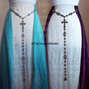 May include: Two cross girdle belts, one silver-toned with blue accents and the other bronze-toned with orange accents. The belts feature a cross pendant and decorative chain. The text "Silver or Bronze Cross Girdle Belt" is visible.