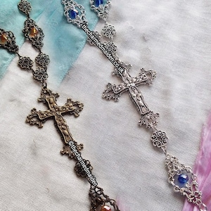 May include: Two ornate rosaries with cross pendants. One is bronze-toned with amber-colored accents, the other is silver-toned with blue accents. Both feature intricate filigree and the text "thecharmedmaiden.co.uk" and "@thecharmedmaiden".