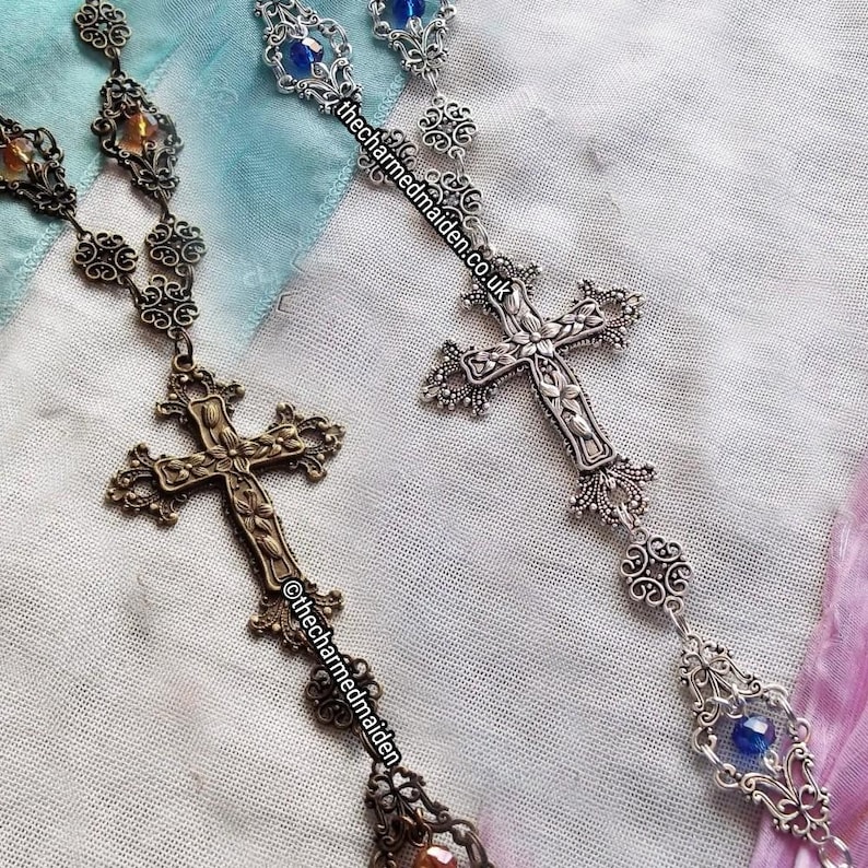 May include: Two ornate rosaries with cross pendants. One is bronze-toned with amber-colored accents, the other is silver-toned with blue accents. Both feature intricate filigree and the text "thecharmedmaiden.co.uk" and "@thecharmedmaiden".