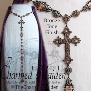 May include: Bronze-toned necklace with a cross pendant and decorative chain. The necklace features a floral design on the cross and is adorned with small, orange-colored beads. The text "The Charmed Maiden" is visible.