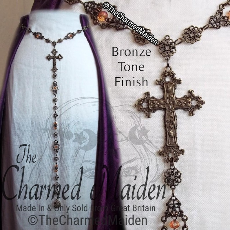May include: Bronze-toned necklace with a cross pendant and decorative chain. The necklace features a floral design on the cross and is adorned with small, orange-colored beads. The text "The Charmed Maiden" is visible.