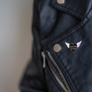 Puede incluir: Una chaqueta de cuero negra con un pin plateado que presenta un coraz&oacute;n negro con alas y el texto "WINGSING IT".