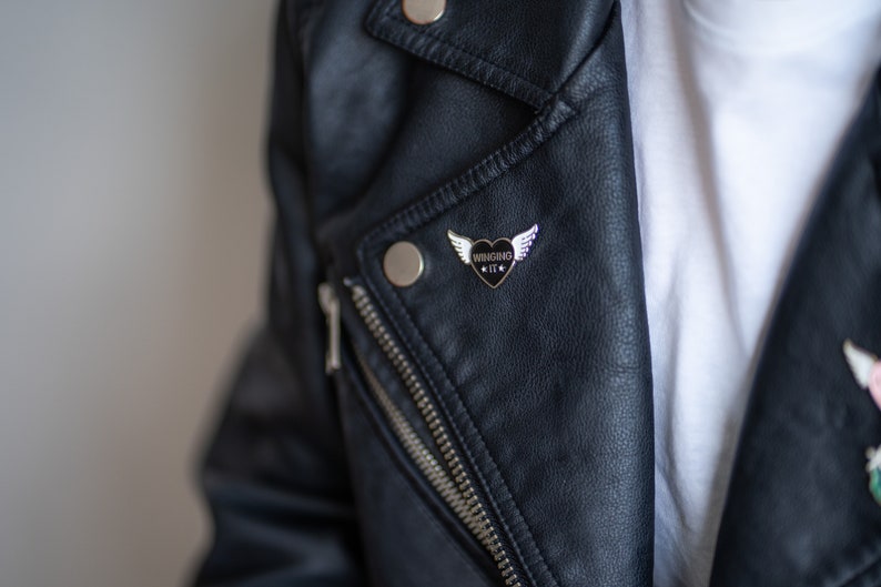 Puede incluir: Una chaqueta de cuero negra con un pin plateado que presenta un coraz&oacute;n negro con alas y el texto "WINGSING IT".