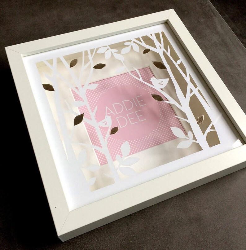 May include: White frame with a white paper cutout design of trees and leaves. The cutout design is layered over a pink background with white polka dots. The text "ADDIE DEE" is in gold lettering on the pink background.