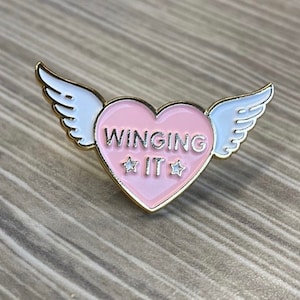 Puede incluir: Un pin de esmalte en forma de coraz&oacute;n rosa con ribete dorado y alas blancas. El coraz&oacute;n tiene el texto "WINGING IT" con dos estrellas blancas a cada lado de la palabra "IT".