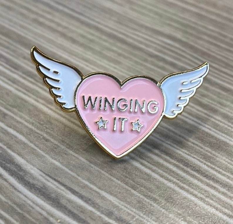 Puede incluir: Un pin de esmalte en forma de coraz&oacute;n rosa con ribete dorado y alas blancas. El coraz&oacute;n tiene el texto "WINGING IT" con dos estrellas blancas a cada lado de la palabra "IT".