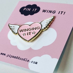 Puede incluir: Un pin de esmalte dorado con un coraz&oacute;n rosa y alas blancas. El coraz&oacute;n tiene el texto "WINGING IT" con dos estrellas a cada lado de la palabra "IT". El pin est&aacute; sobre un fondo rosa con una nube blanca y el texto "PIN IT WING IT!" y "www.pipanddoodle.com".