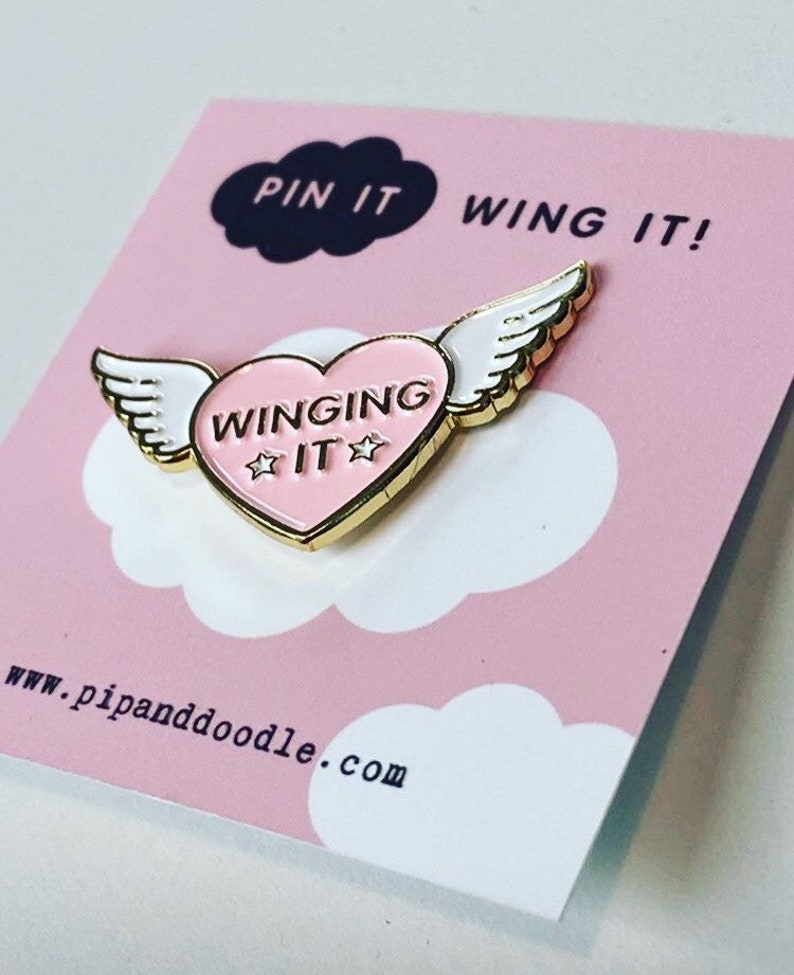 Puede incluir: Un pin de esmalte dorado con un coraz&oacute;n rosa y alas blancas. El coraz&oacute;n tiene el texto "WINGING IT" con dos estrellas a cada lado de la palabra "IT". El pin est&aacute; sobre un fondo rosa con una nube blanca y el texto "PIN IT WING IT!" y "www.pipanddoodle.com".