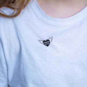 Puede incluir: Una camiseta blanca con un pin de esmalte negro que representa un coraz&oacute;n con alas y el texto "WINGING IT".