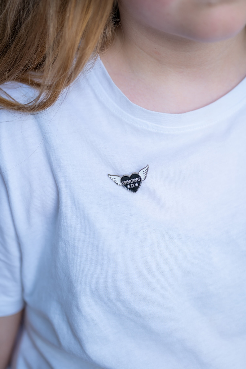 Puede incluir: Una camiseta blanca con un pin de esmalte negro que representa un coraz&oacute;n con alas y el texto "WINGING IT".