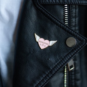 Puede incluir: Un pin de esmalte en forma de coraz&oacute;n rosa con alas blancas y el texto "WINGING IT" en una chaqueta de cuero negra.