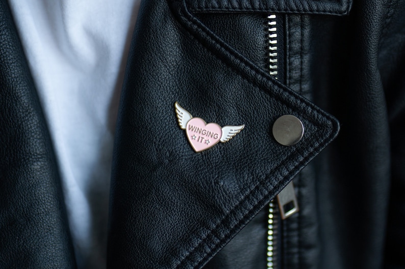 Puede incluir: Un pin de esmalte en forma de coraz&oacute;n rosa con alas blancas y el texto "WINGING IT" en una chaqueta de cuero negra.
