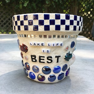 Puede incluir: Maceta de cerámica blanca con un patrón de cuadros azules y blancos alrededor de la parte superior. La maceta está decorada con azulejos de mosaico azules y las palabras "Lake Life is Best" en letras negras.