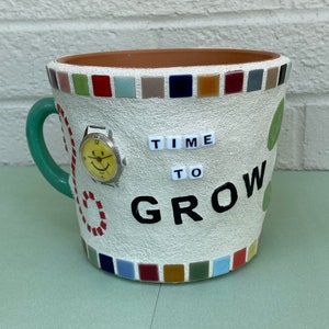 Könnte beinhalten: Weißer Keramiktopf mit grünem Griff, verziert mit bunten Mosaikfliesen, einer Vintage-Uhr und den Worten "TIME TO GROW".