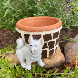 Puede incluir: Una pequeña figurita de perro de cerámica blanca se encuentra junto a una maceta de mosaico marrón y blanco. La maceta está hecha de terracota y tiene un esmalte marrón. El perro tiene un cuerpo blanco y ojos negros.