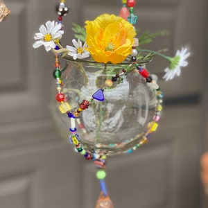 May include: A glass vase with a yellow flower and white flowers hangs from a colorful beaded chain with a bell at the bottom.